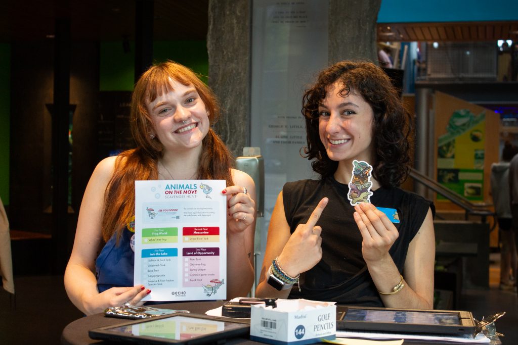 ECHO Staff members Ava and Colyar smiling at the welcome table of an event while holding up a scavenger hunt and stickers.