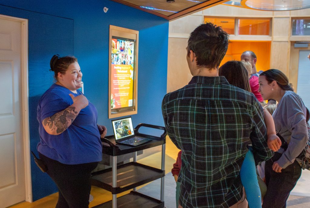 Image of ECHO Animal Care specialist Nicole Dawson pointing over her shoulder at the Into the Lake gallery, currently closed for construction, as she speaks to a group of guests. Next to Nicole a laptop displays behind-the-scenes images of ECHO's renovation process.