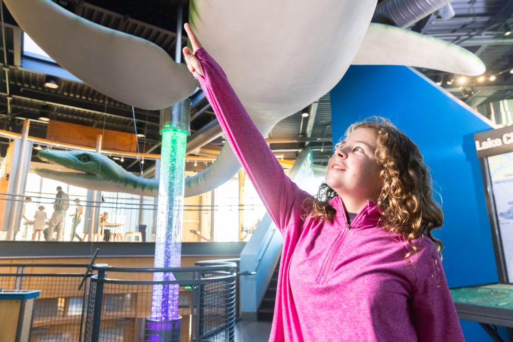 A child in a pink zip-up sweater points upward at a giant plesiosaur sculpture.