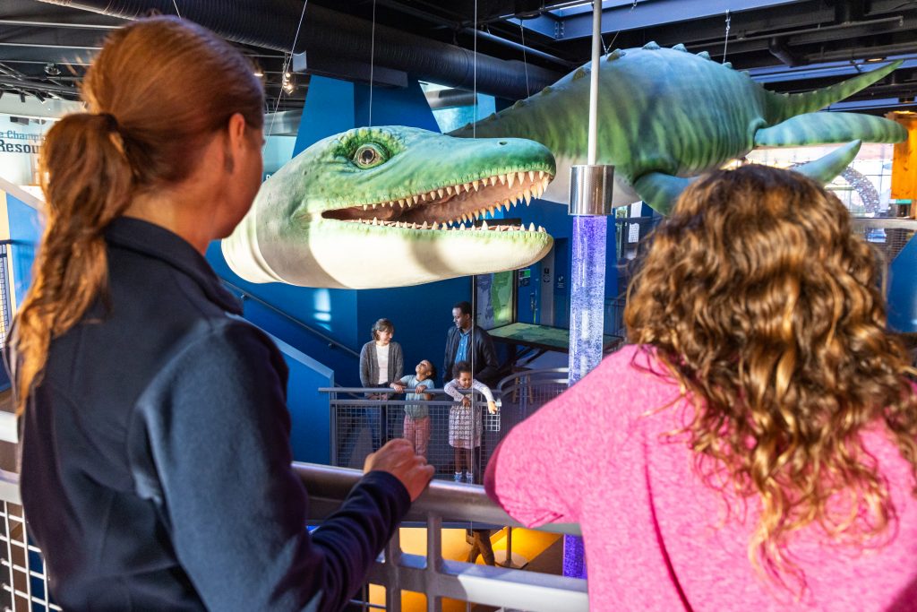 An adult and child stand at a railing and face away from the camera, towards ECHO's 30 foot Champ sculpture. A family is visible on the floor below looking upward at Champ.