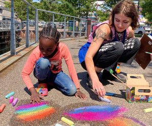 An ECHO Camp counselor and camper crouch down to draw on the pavement with colorful chalk