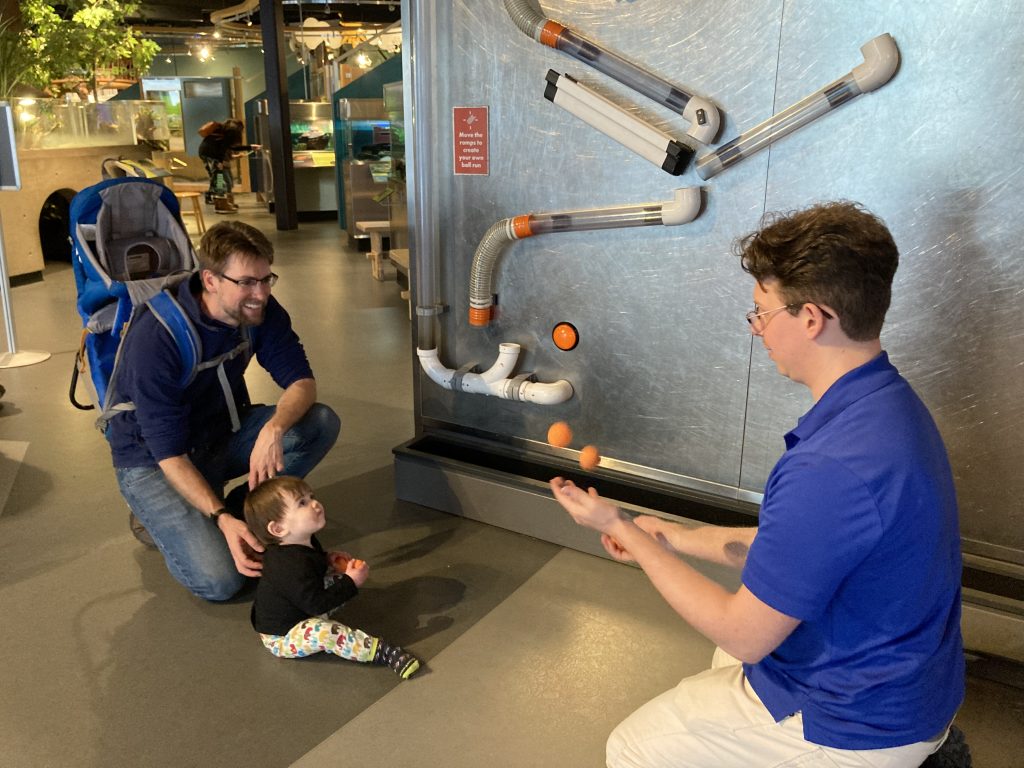 Photo of a dad smiling with young child in front of the ball wall exhibit. Museum employee is juggling with the balls.