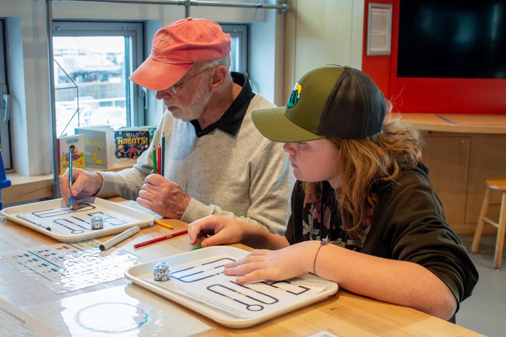 An adult and child sit inside ECHO's Engineer It space. In front of them on the table are two trays with paper, colored pencils, and small round robots.