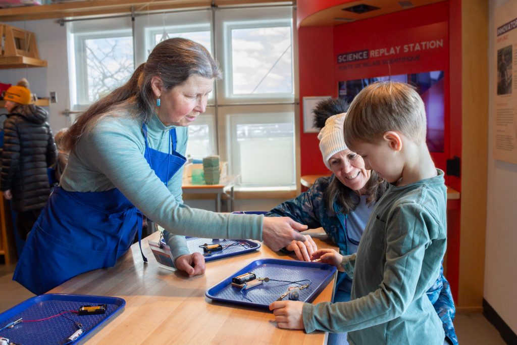 A parent, child, and ECHO volunteer work together to build a circuit using wires, a battery pack, and a motor inside ECHO's Engineer It activity space.