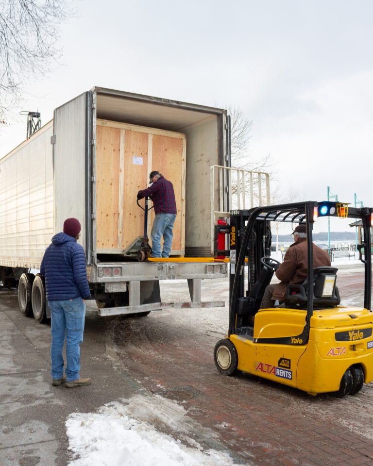 An ECHO employee watches as ECHO Facilities and Animal Care director Steve sits in a forklift waiting to remove a large wooden crate from a semi truck. Inside the trailer of the truck, a delivery driver uses a pallet jack to pull the crate towards the trailer doors.