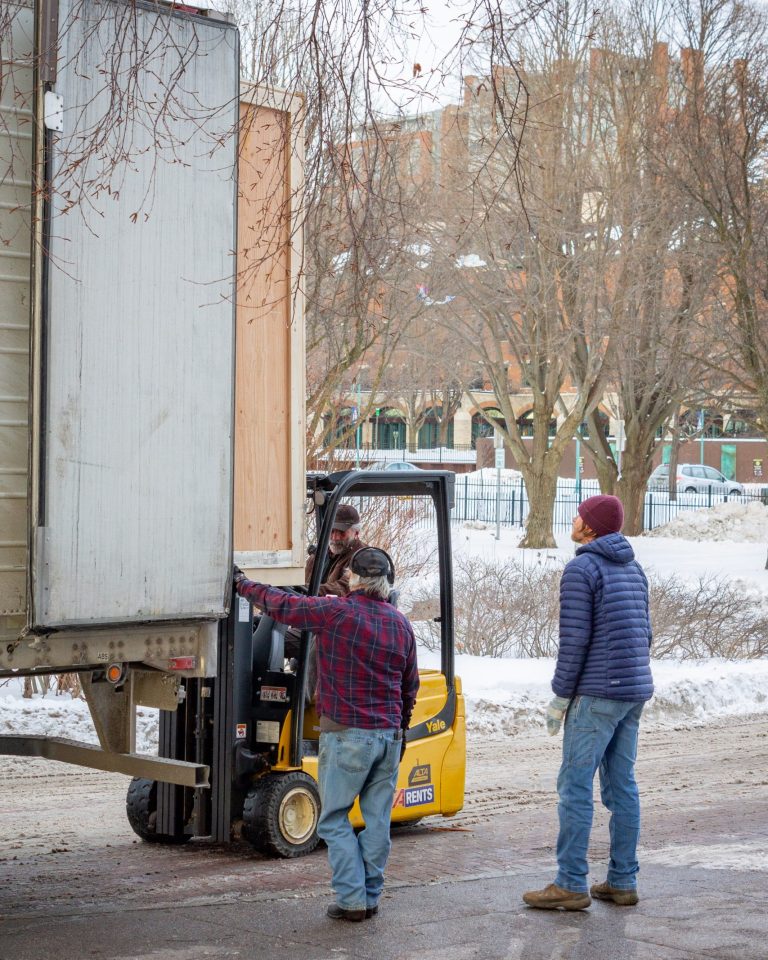 Two adults watch as ECHO Facilities and Animal Care Director Steve uses a forklift to lift a massive wooden crate out of the back of a semi truck. The crate is nearly as tall as the trailer.