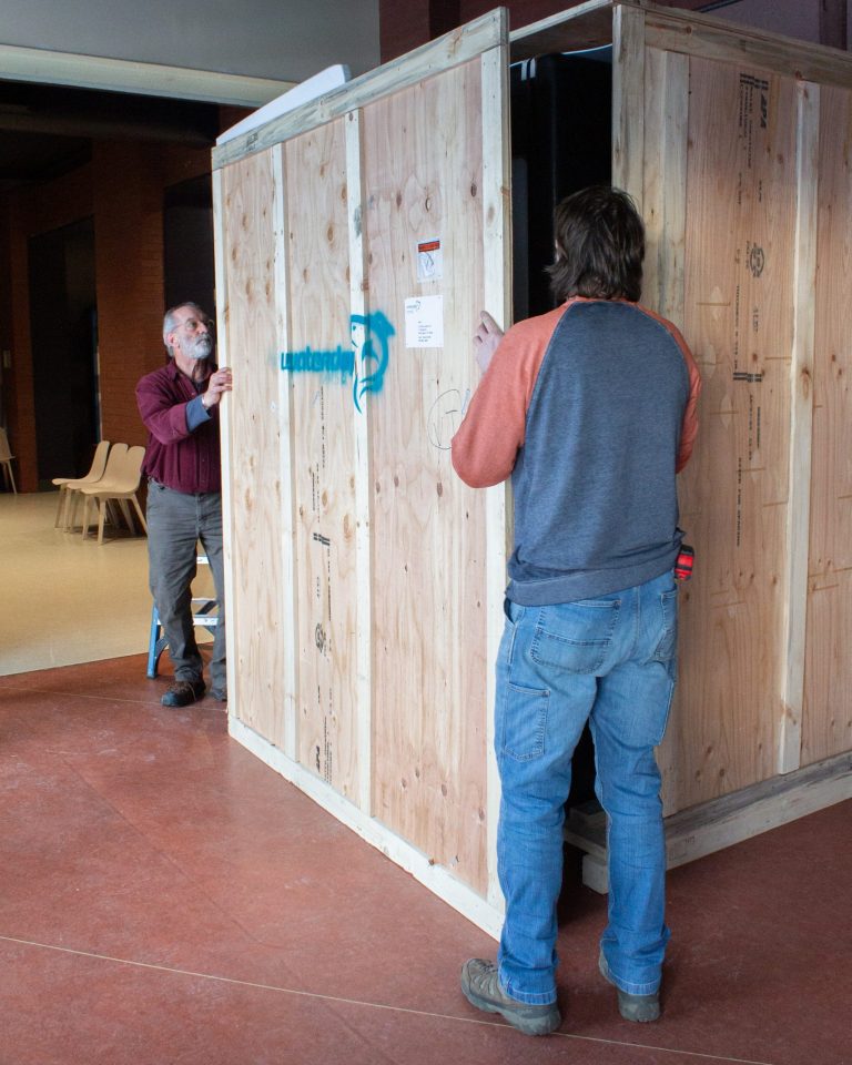 Two ECHO staff members remove the front panel from a massive wooden crate. The crate is taller than both the employees and is so large it extends out of view.